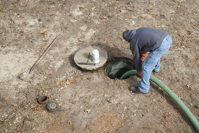 Worker in a gray hoodie and orange gloves feeding a green suction hose into an open septic tank in a leaf-strewn yard, the round concrete lid lying beside it