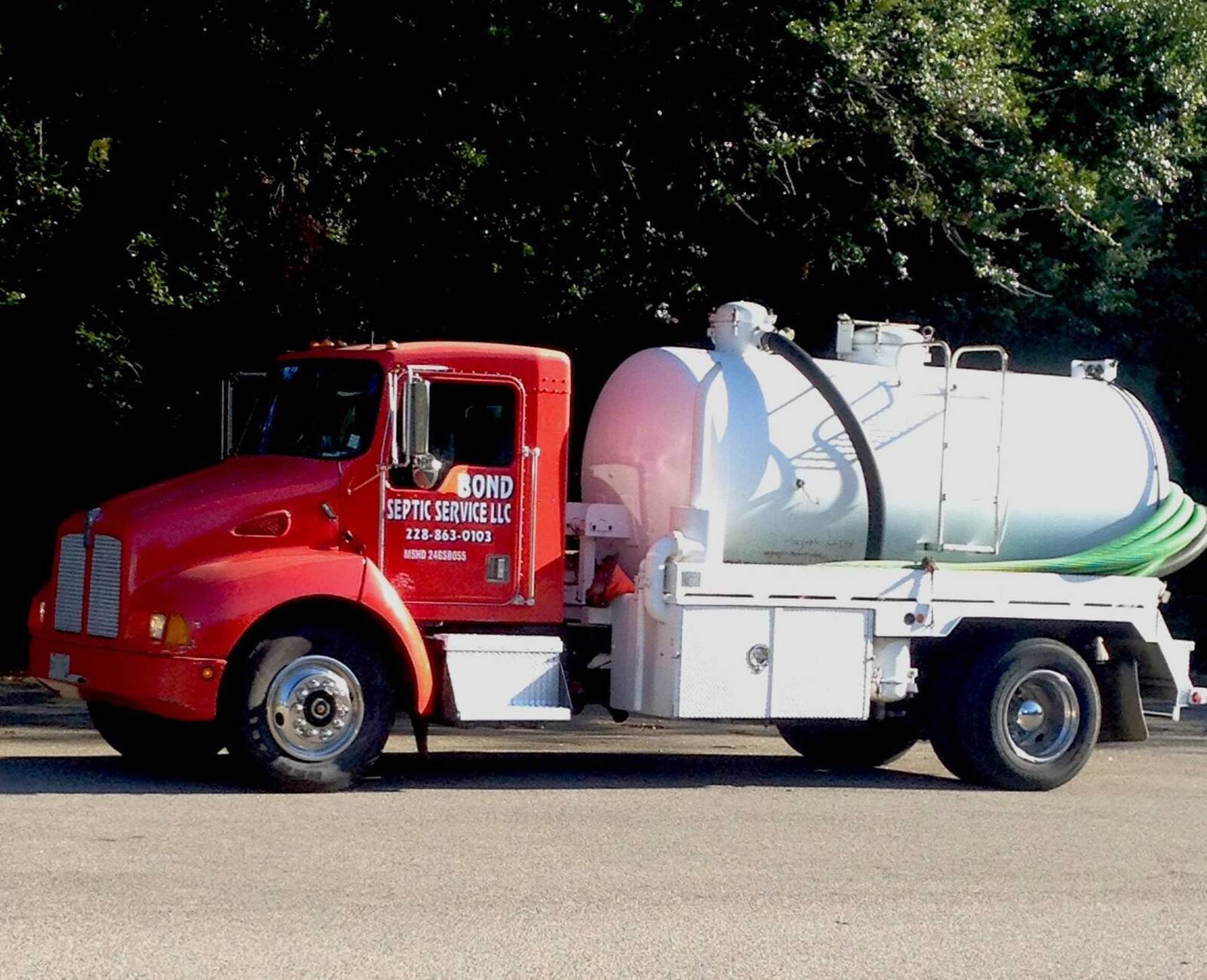 Red truck with a white vacuum tank and a coiled green suction hose, BOND SEPTIC SERVICE LLC and 228-863-0103 lettered on the door, parked in front of dark trees
