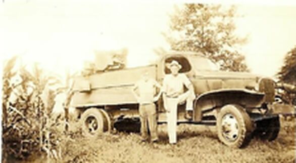 Old sepia photograph of two men, one in a wide-brim hat, standing beside an old flatbed truck next to a row of corn